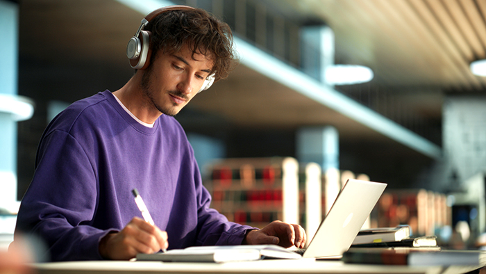 A student with headphones makes notes as he works on his laptop in a library.