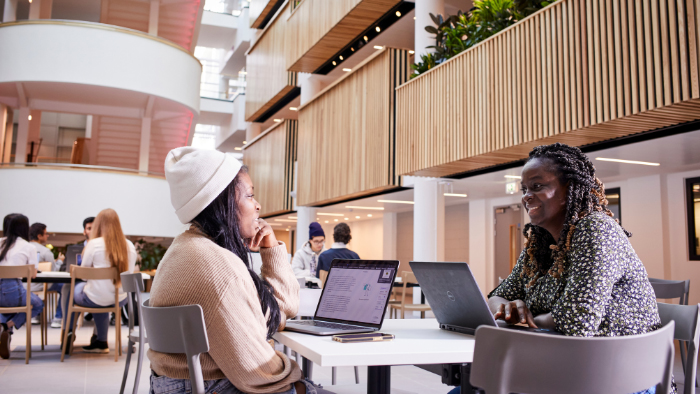 Two students talking in The Atrium, laptops in front of them.