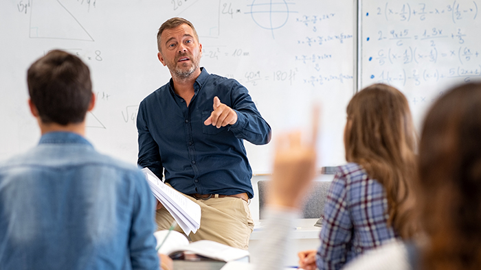 A teacher in a bright modern classroom. A student is volunteering to answer a question.