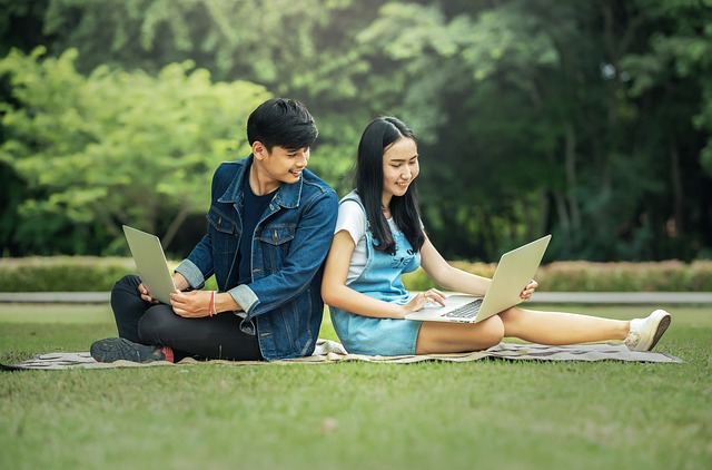 Image of 2 students sitting in a park working on their laptops