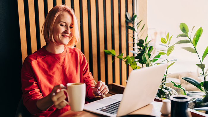 A young adult working on a laptop, smiling and drinking coffee. 