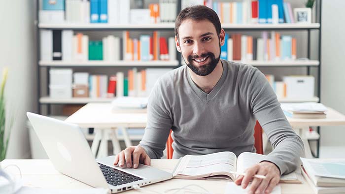 a smling student is working at an open laptop with a pile of books infront of him