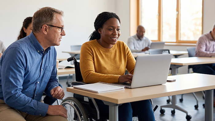 A helpful teacher, is sat next to a student in a wheelchair as they look at a laptop together.