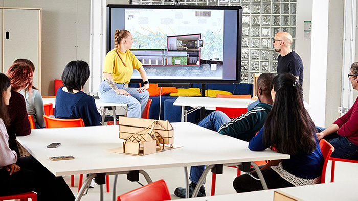 A classroom, with a young adult presenting to an engaged audience as an older mentor looks on.