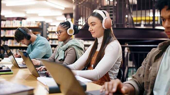a group of students are working individually in a library. They are wearing large headphones as they study.