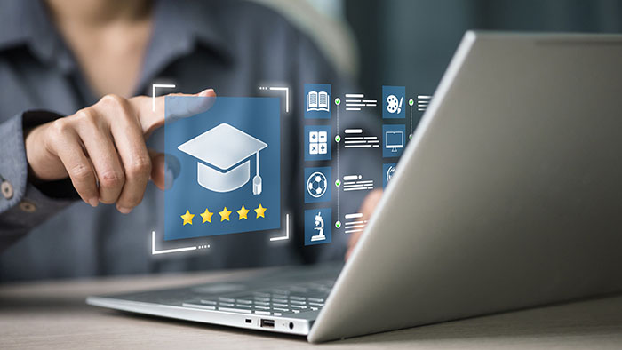 An open laptop has some virtual educational icons hovering above the keyboard. An unseen user is selecting the mortar board. 