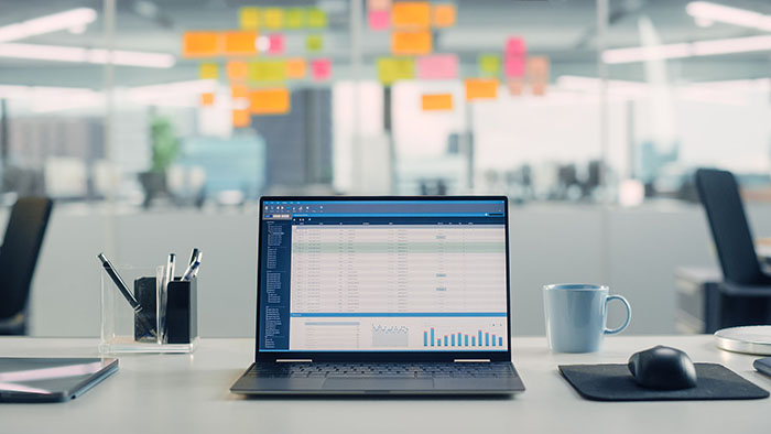 An open laptop is on a desk in a well-lit office. There are post-it notes on the glass wall behind the computer.