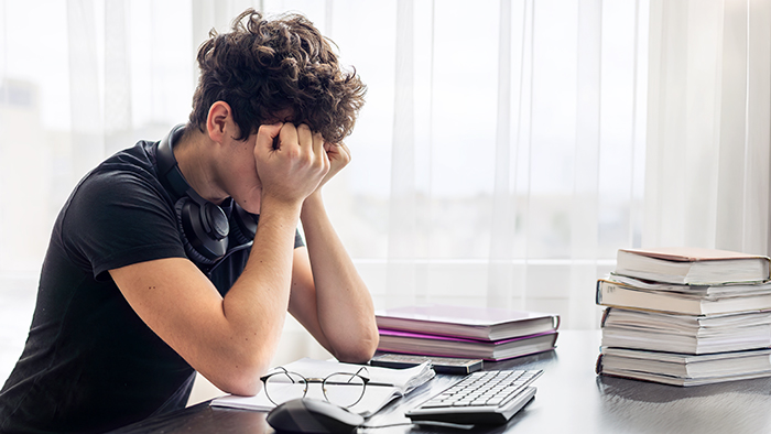 A male student sat alone at a desk, surrounded by piles of books, with their head in their hands.