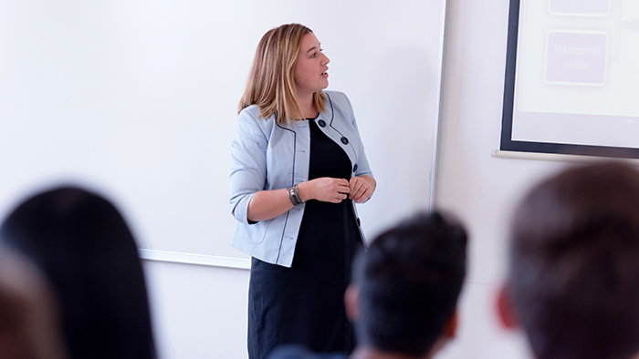 A woman by a whiteboard, presenting to a small group of adults.