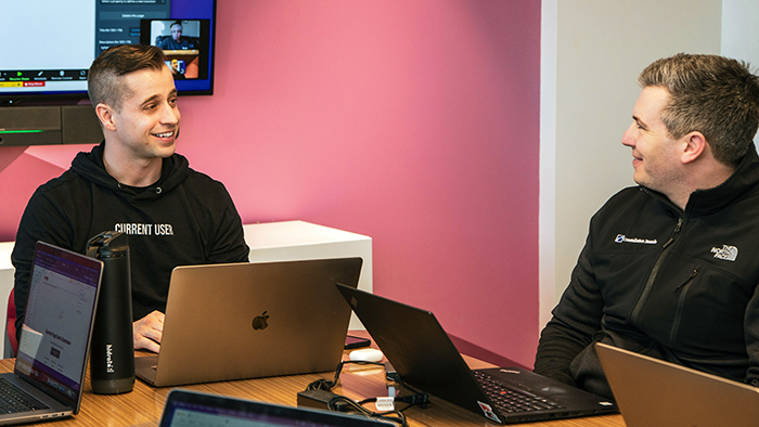 Two young academics chatting working on laptops and chatting with each other.